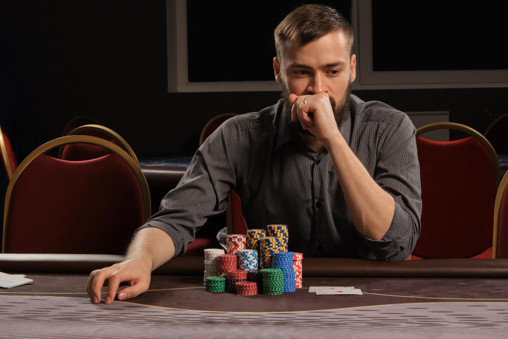 Handsome bearded male in a gray shirt is playing poker sitting at the table